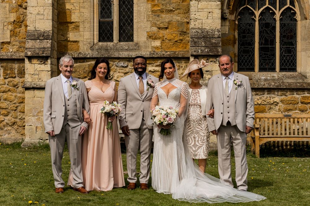 Wedding party posing outside a historic stone church, featuring bride, groom, and family in elegant formal attire.