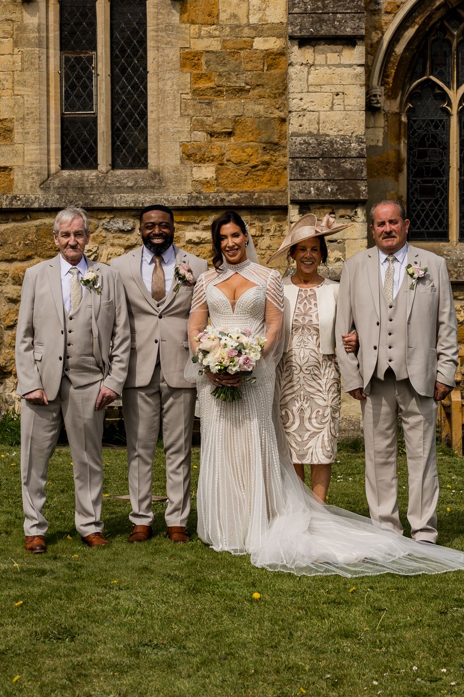 Wedding party posing outside stone church, bride in white gown with bouquet, groom and groomsmen in beige suits.