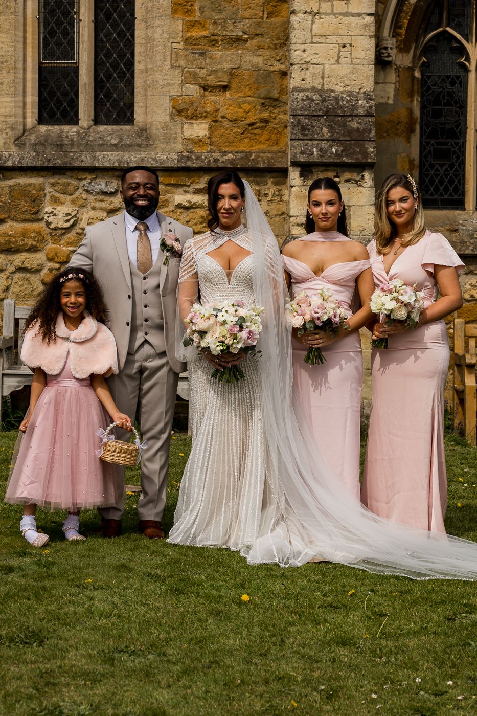 Bride in white gown with bridesmaids in pink dresses and groom in grey suit posing outside a stone church.