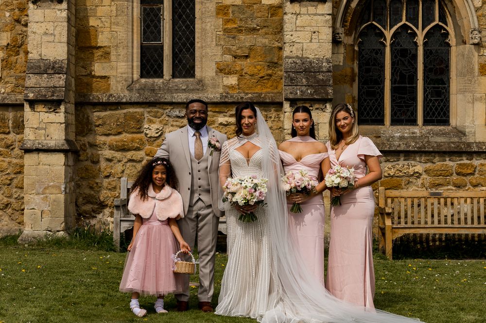 Wedding party poses outside stone church, bride in white gown, groom in gray suit, bridesmaids in pink, flower girl.