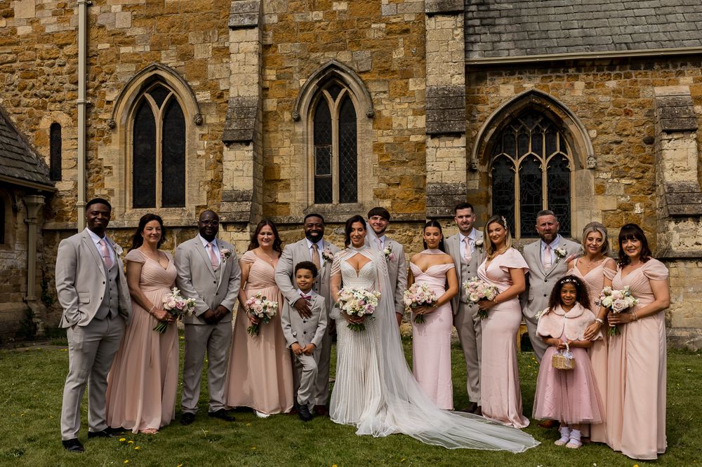 Wedding party posing outside a stone church, bride in white gown with bridesmaids in blush pink dresses and groomsmen in grey suits.