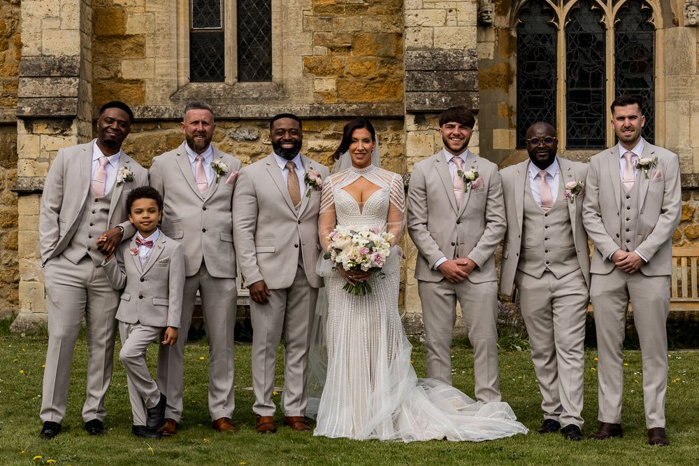 Bride and groom pose with groomsmen in tan suits outside a stone church on their wedding day.