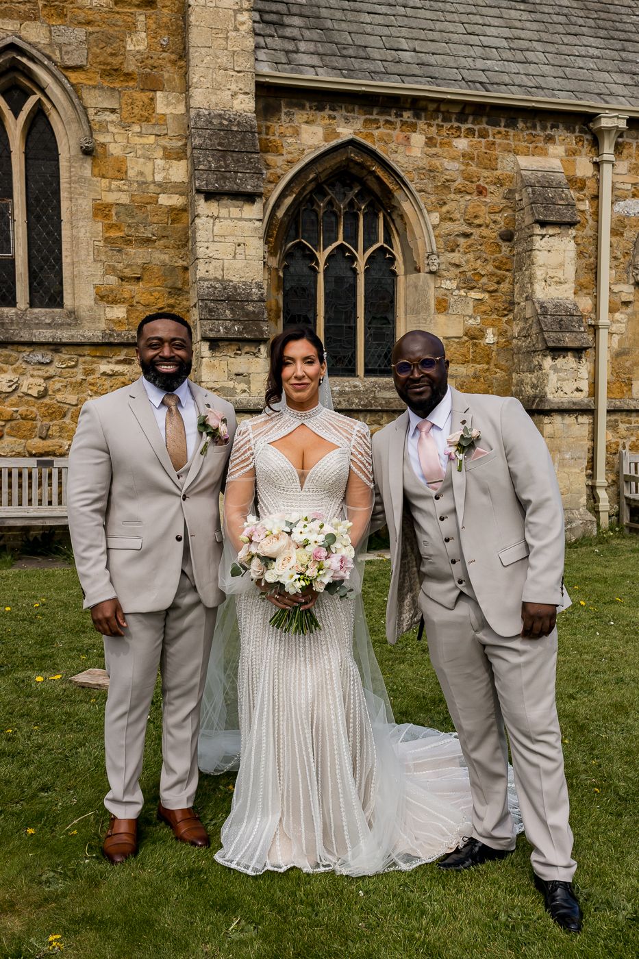Bride in beaded gown holding bouquet poses with two groomsmen in tan suits outside a stone church.