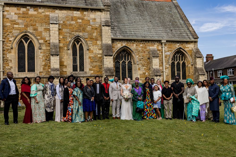 Large wedding group photo outside a historic stone church, guests dressed in colorful African attire with bride and groom centered.