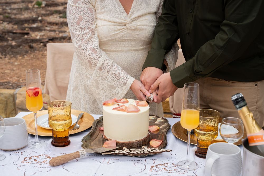 Couple cutting a white wedding cake topped with peaches at an outdoor reception table with champagne glasses.
