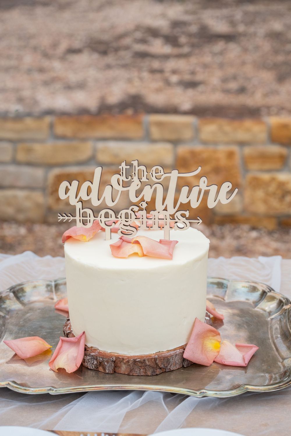 White wedding cake with adventure begins topper on rustic wood slice surrounded by pink rose petals.