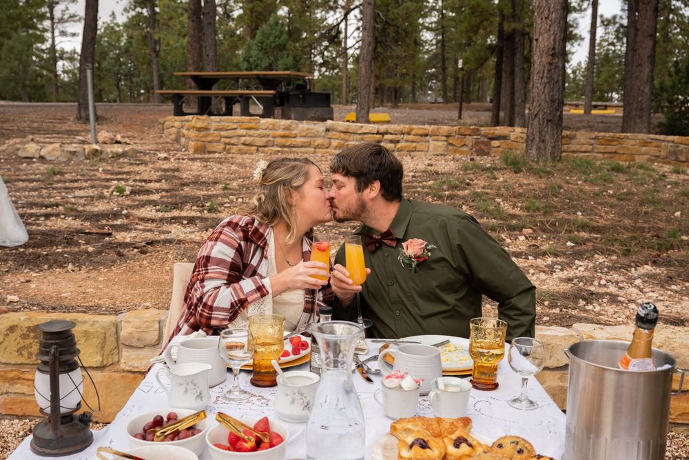 Couple kissing over mimosas at an outdoor breakfast table in a forest setting.