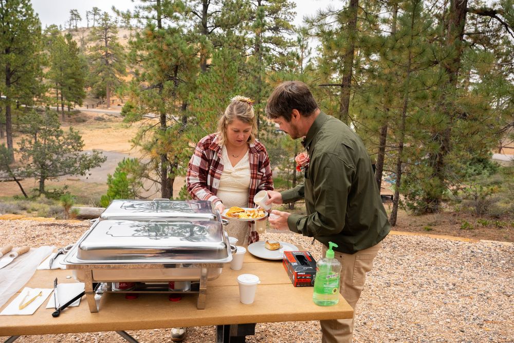 Couple serving food at an outdoor picnic table with a chafing dish, surrounded by pine trees in a natural setting.
