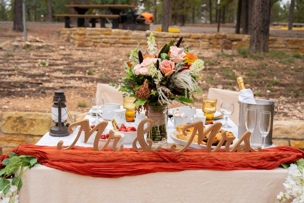 Rustic outdoor wedding sweetheart table with Mr. and Mrs. signs, floral centerpiece, and red table runner in a forest setting.