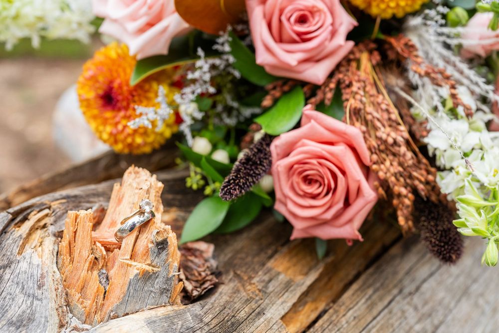 Pink roses and colorful wildflowers arranged on a rustic wood stump with wedding rings nearby.