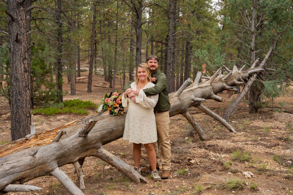 Couple posing in a pine forest beside a fallen log, woman holding a bouquet, man embracing her from behind.