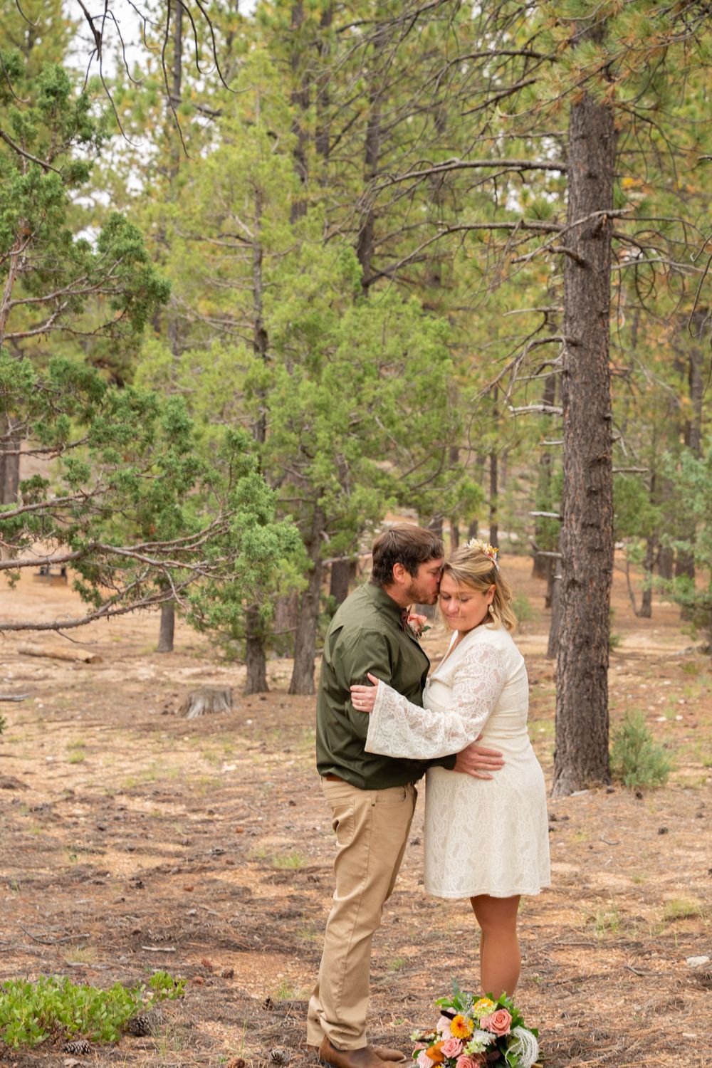 Couple embracing in a pine forest during an outdoor wedding ceremony, bride in white dress holding a colorful bouquet.