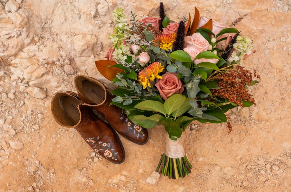 Rustic bridal bouquet with pink roses and wildflowers beside brown leather cowboy boots on a sandy stone surface.