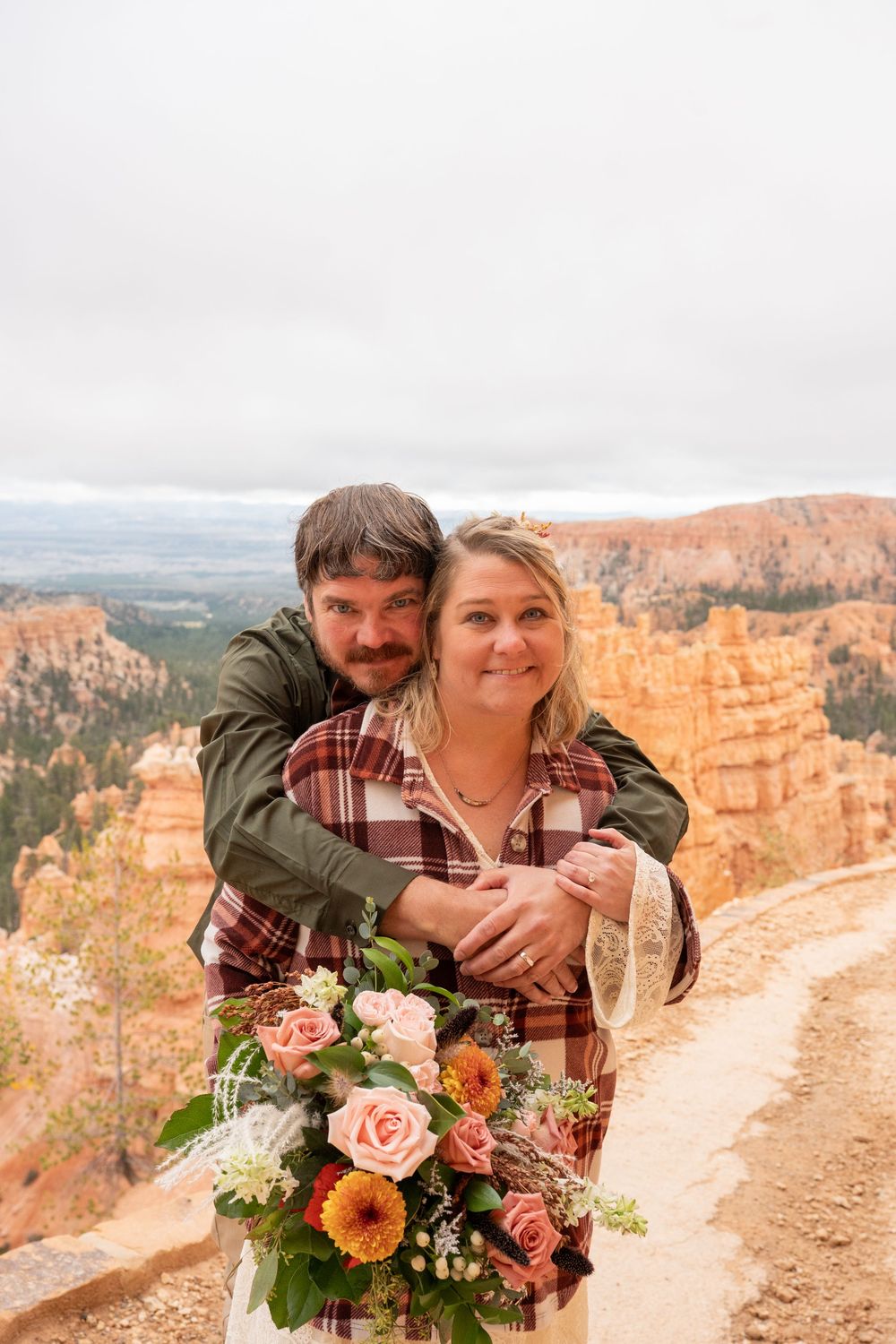 Happy couple embraces at Bryce Canyon overlook, bride holding colorful bouquet of roses and gerbera daisies.