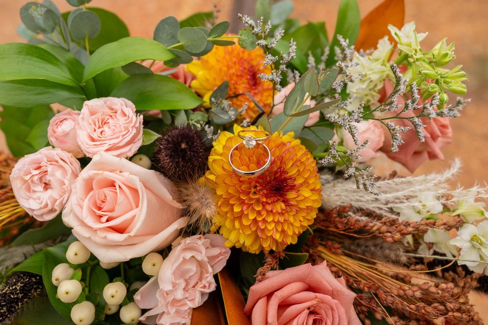 Wedding rings rest on a lush bouquet of pink roses, orange dahlias, and wildflowers in warm autumn tones.