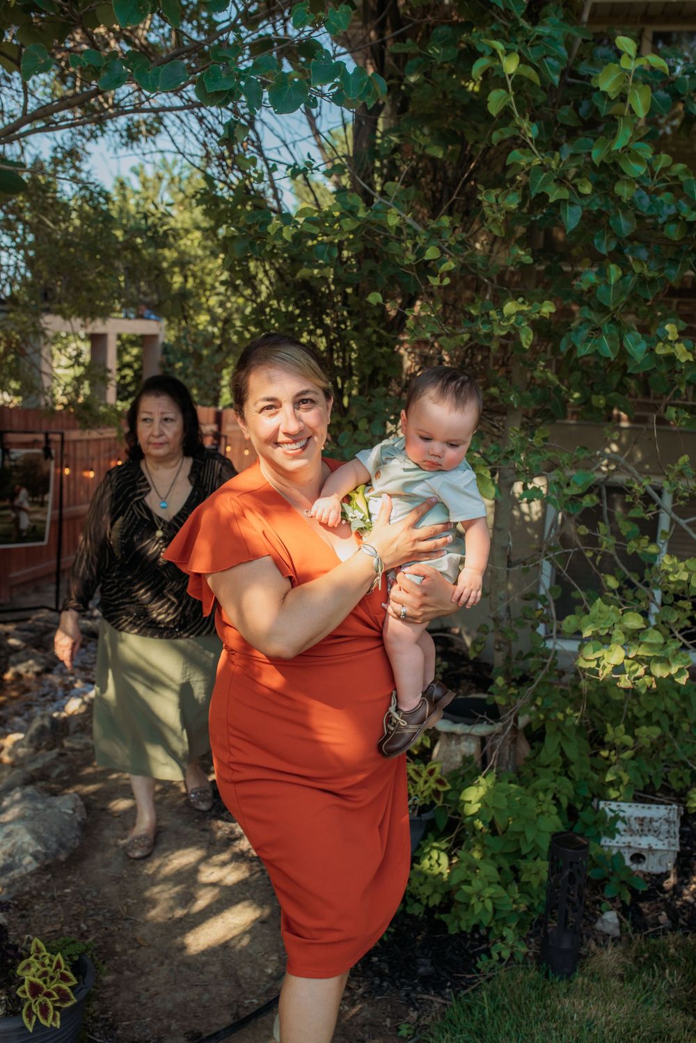 Woman in orange dress holds baby in lush garden setting with older woman walking behind her.