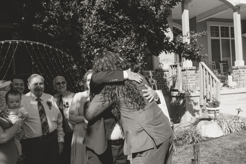 Black and white wedding photo of couple embracing outdoors, guests watching in background near decorated venue.