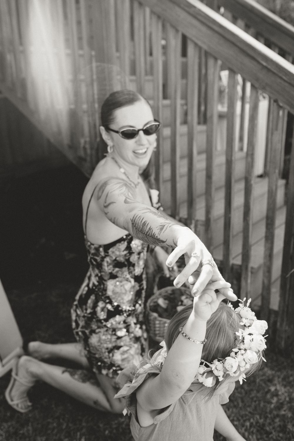 Black and white photo of smiling woman in sunglasses placing floral crown on young girls head outdoors.