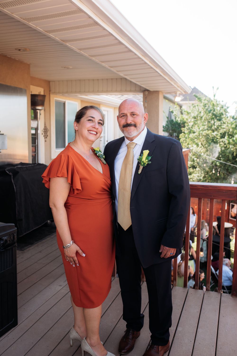 Elegant couple posing on deck at wedding, woman in rust orange dress, man in navy suit with boutonniere.