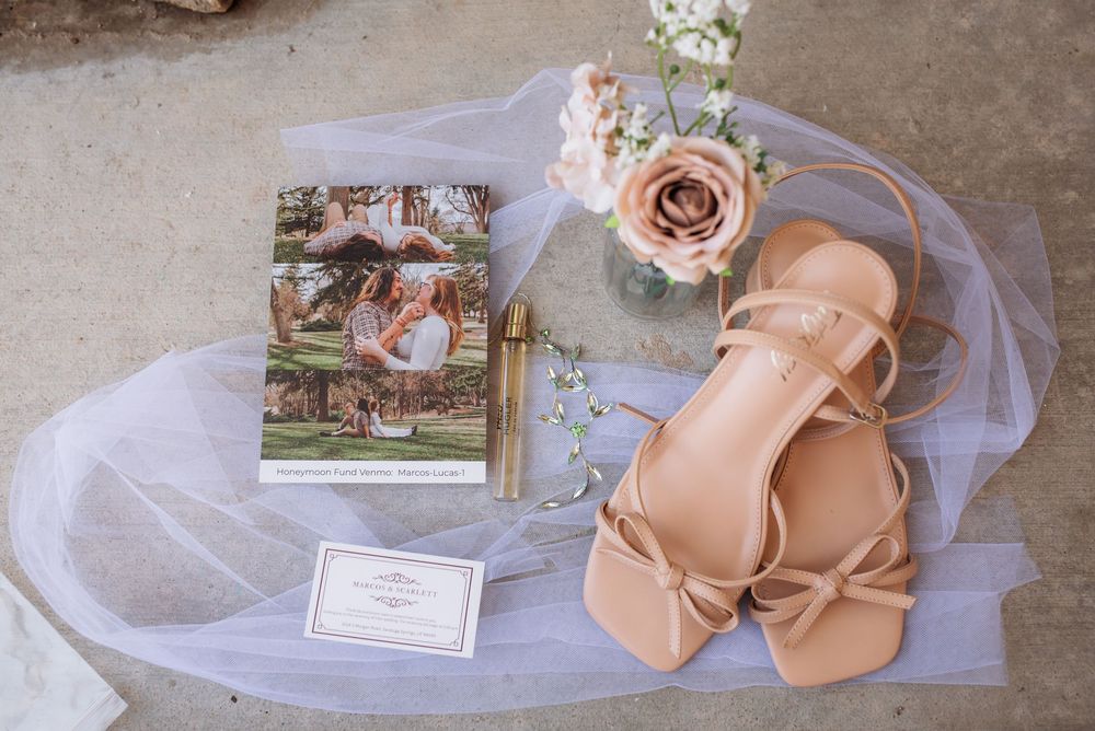 Bridal flat sandals beside a wedding program, veil, and pink roses on a neutral background.