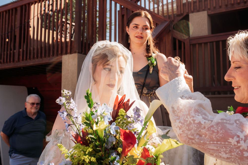 Bride in white gown and veil holds colorful bouquet while bridesmaids assist during outdoor wedding ceremony.