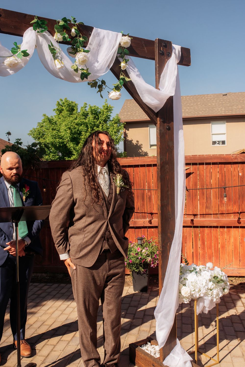 Groom in brown suit stands under floral wedding arch with white draping at outdoor backyard ceremony.