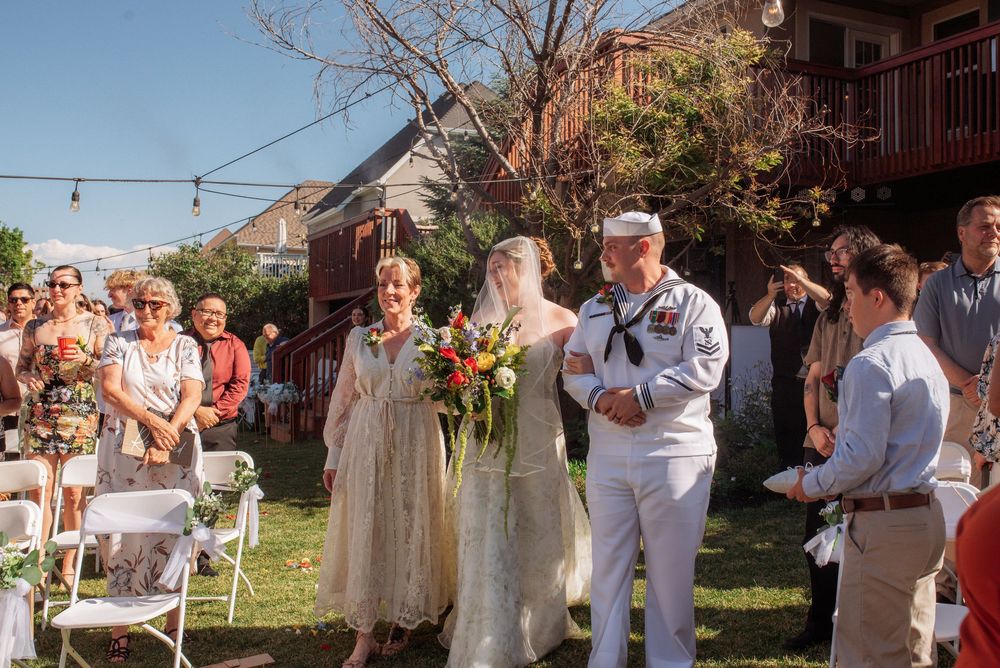 Bride in white gown walks down outdoor aisle with Navy sailor and woman at sunny backyard wedding ceremony.