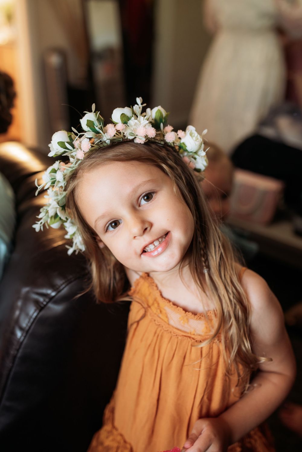 Young girl in orange dress wearing a floral crown of white and pink roses smiles sweetly indoors.