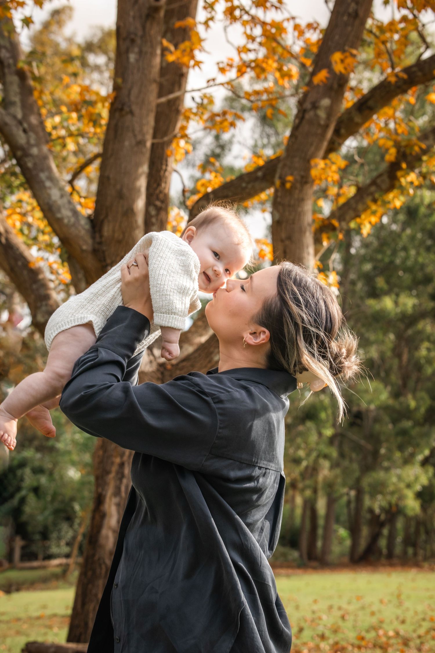 A mother in a dark button-up shirt lifts and kisses her smiling baby under autumn trees with golden leaves in a sunlit park in Castle Hill.