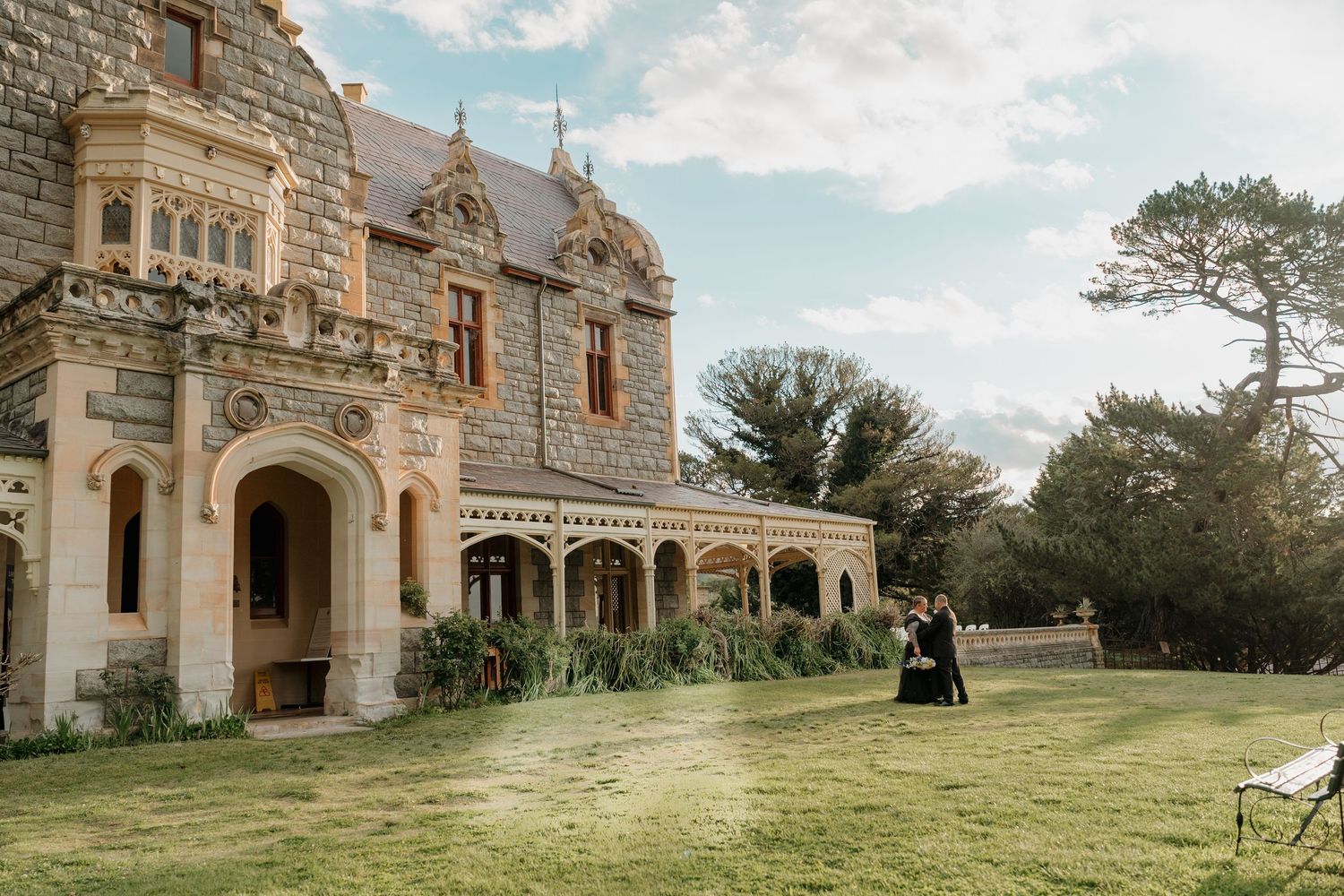 A couple in Victorian attire poses on the lawn of a grand Gothic stone manor under a cloudy sky.