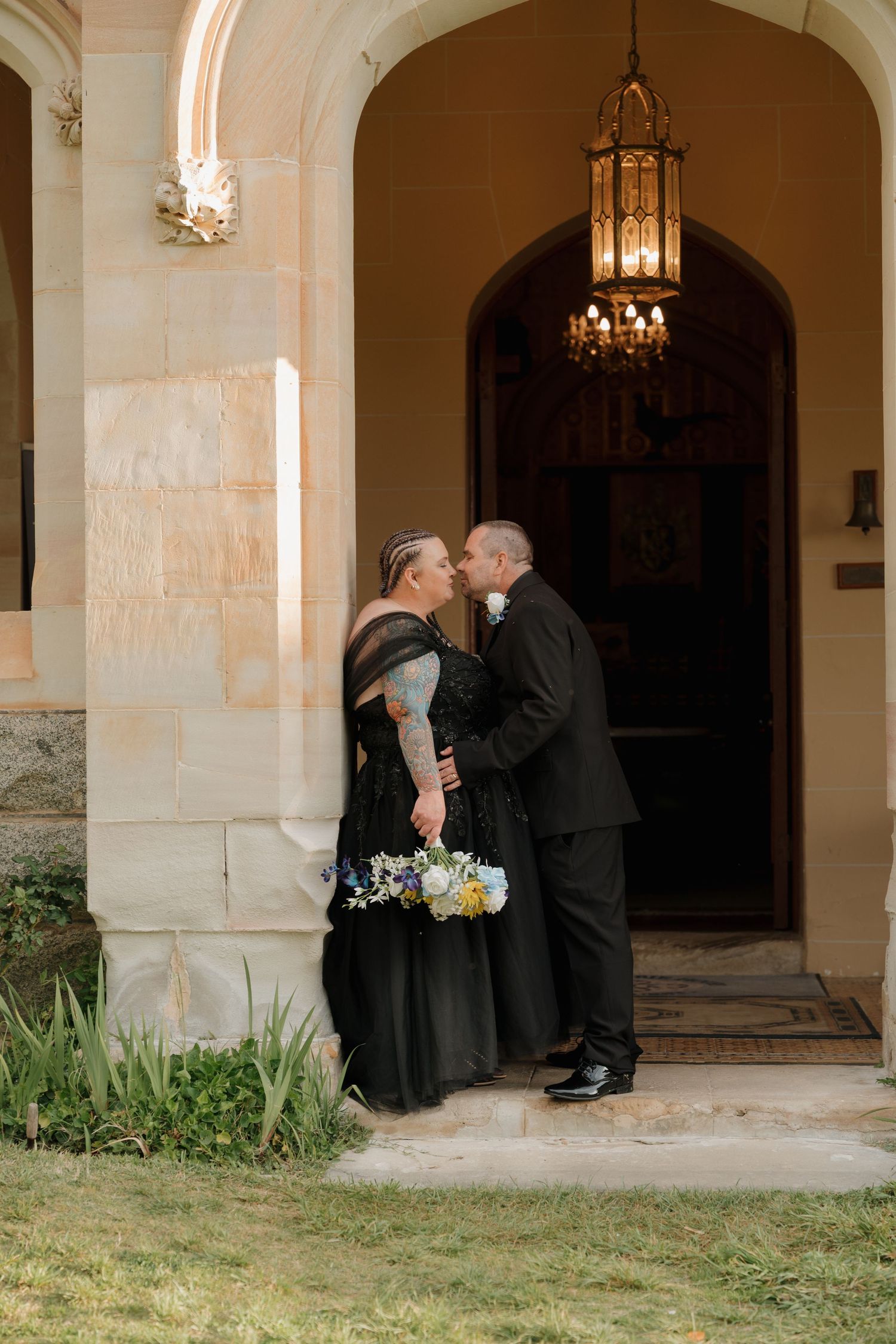 Couple in black wedding attire sharing a kiss outside an elegant stone arch entrance with chandelier.