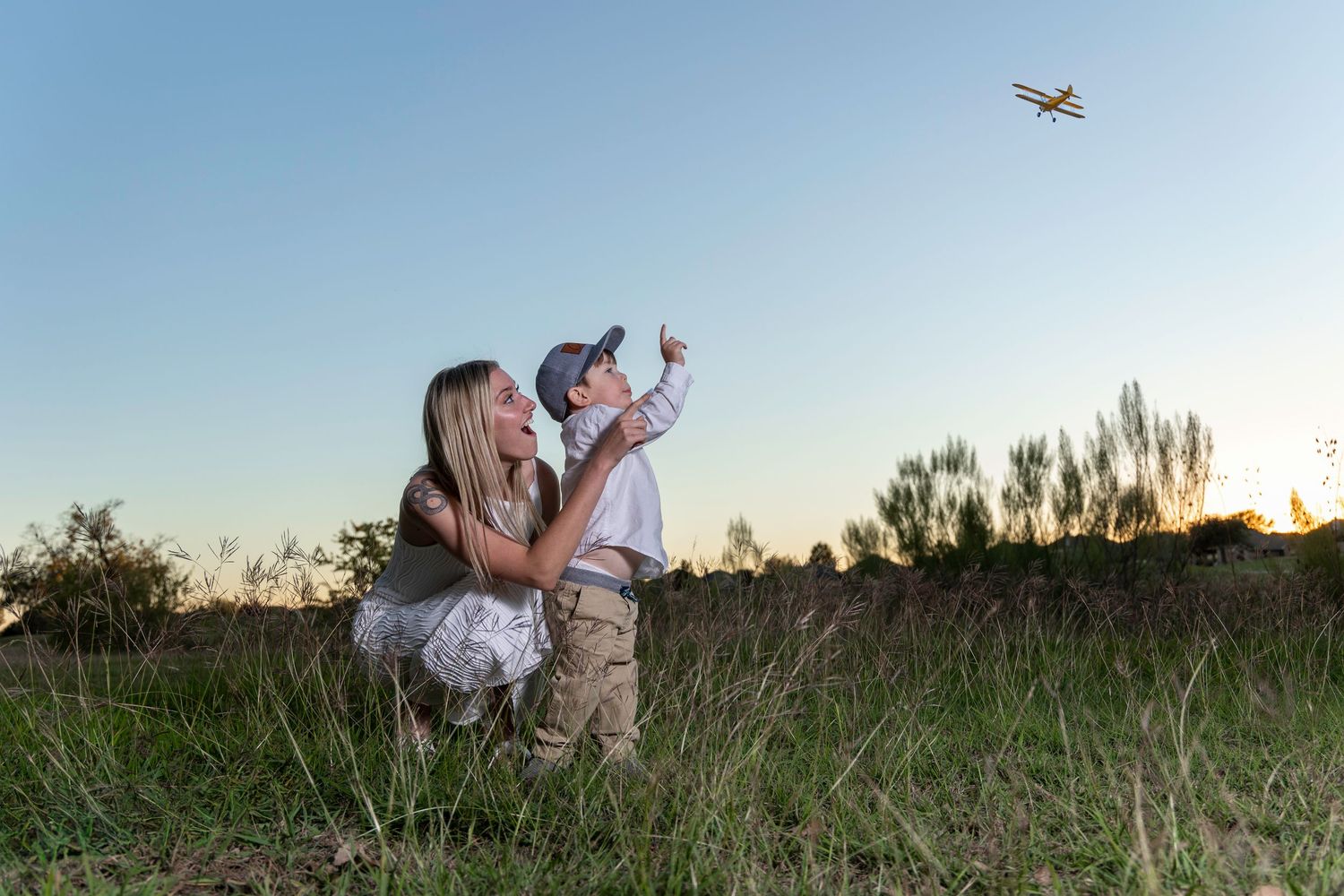 A mother and young boy playing with a toy airplane in a grassy field at sunset.