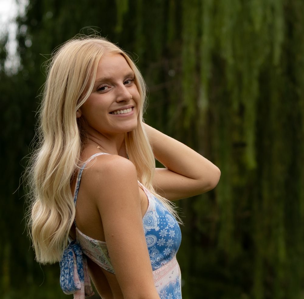 Young blonde woman smiling outdoors in a floral dress with lush green weeping willow trees in the background.
