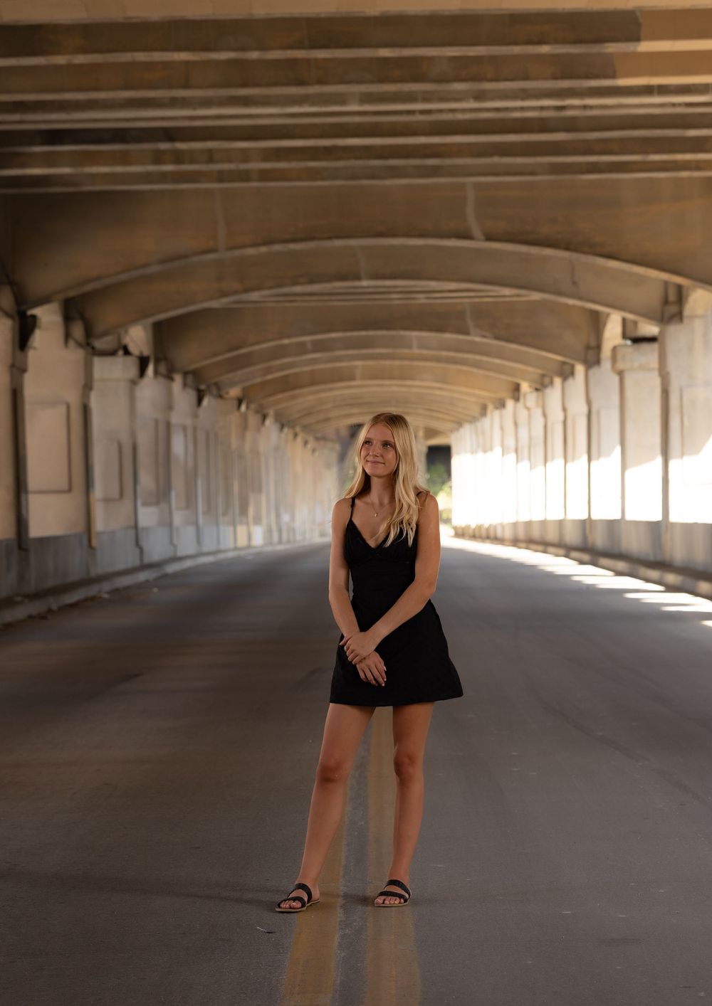 Blonde woman in black mini dress and sandals standing under an arched bridge tunnel with golden backlight.
