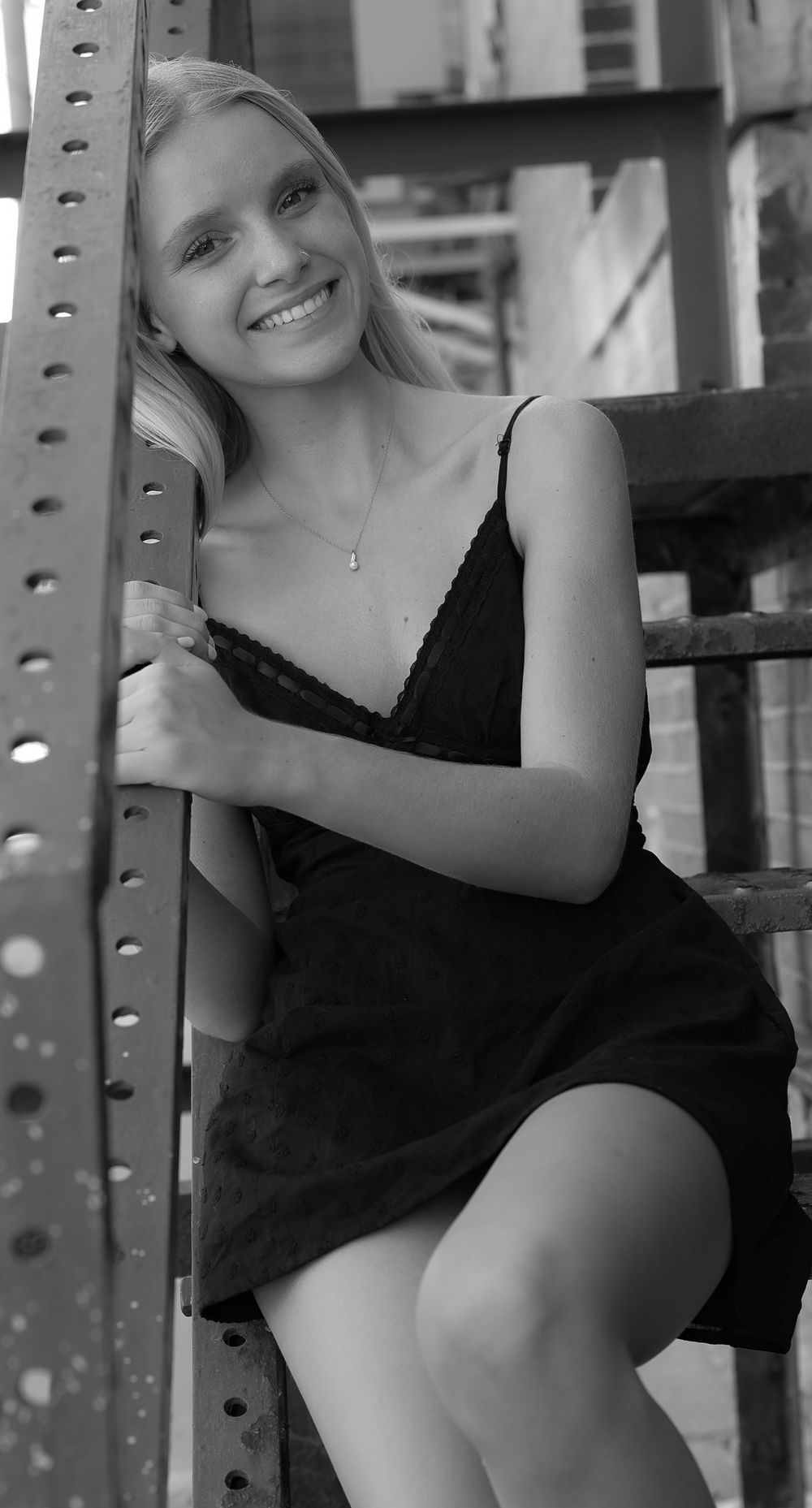 Smiling young woman in black slip dress posing against metal structure in black and white photo.