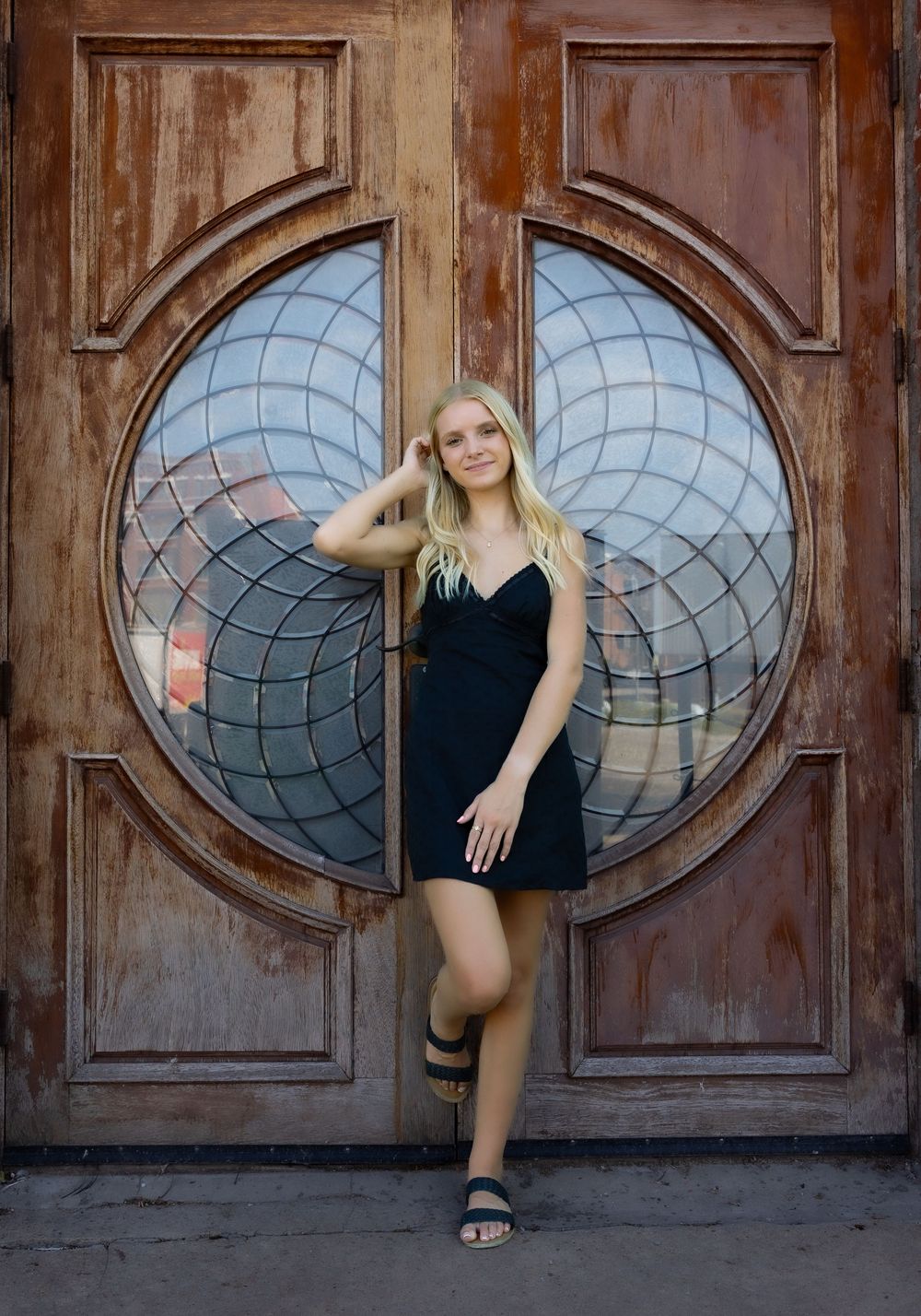 Young blonde woman in black dress posing in front of ornate wooden doors with circular stained glass window.