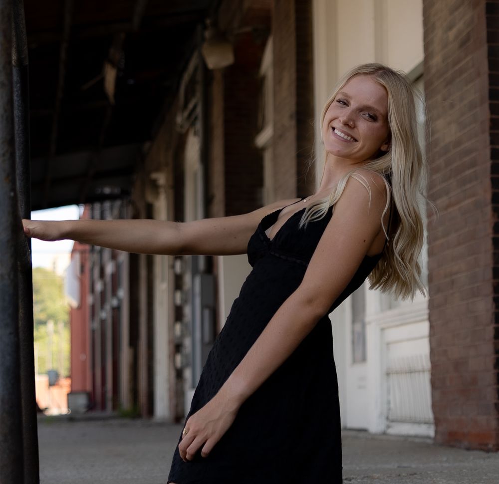 Young blonde woman in black dress posing outdoors near brick building columns, smiling naturally.