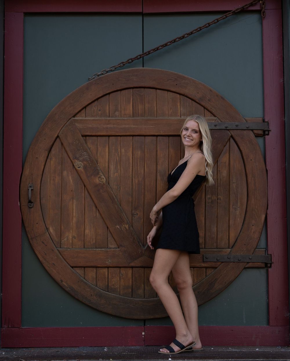 Smiling blonde woman in black dress posing in front of large circular wooden barn door.