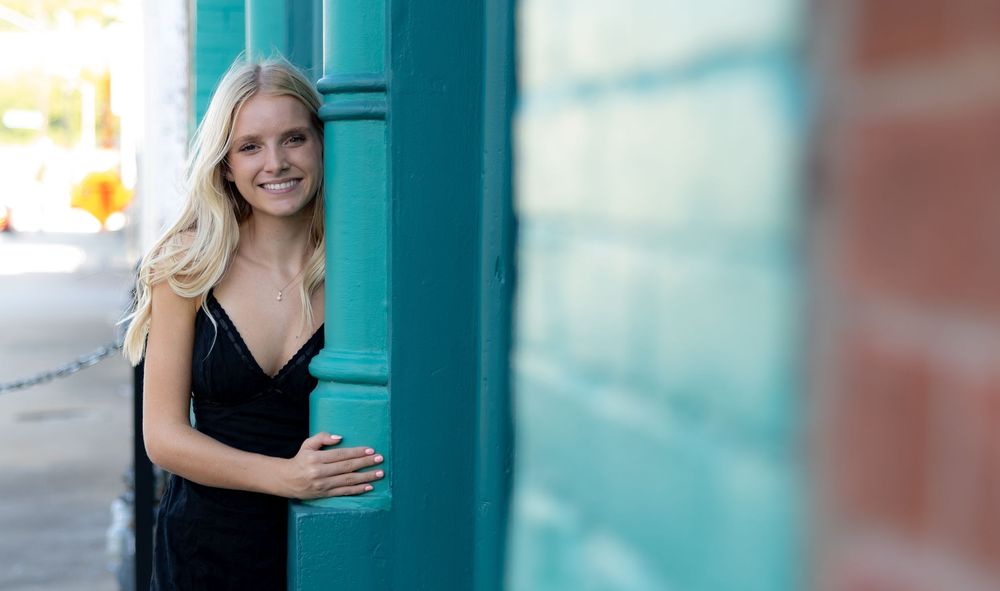Young blonde woman in black dress posing by teal door in urban outdoor setting.