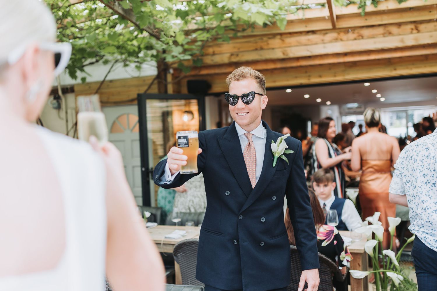 Groom in navy suit and sunglasses raises a glass at outdoor wedding reception venue.