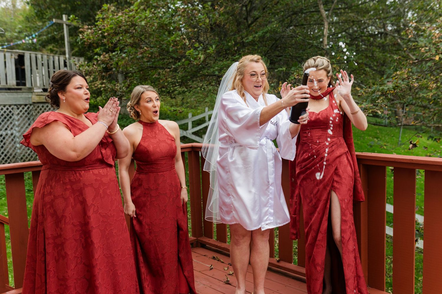Bride in a white robe and veil reacts excitedly on a wooden deck as three bridesmaids in red dresses cheer and clap around her.
