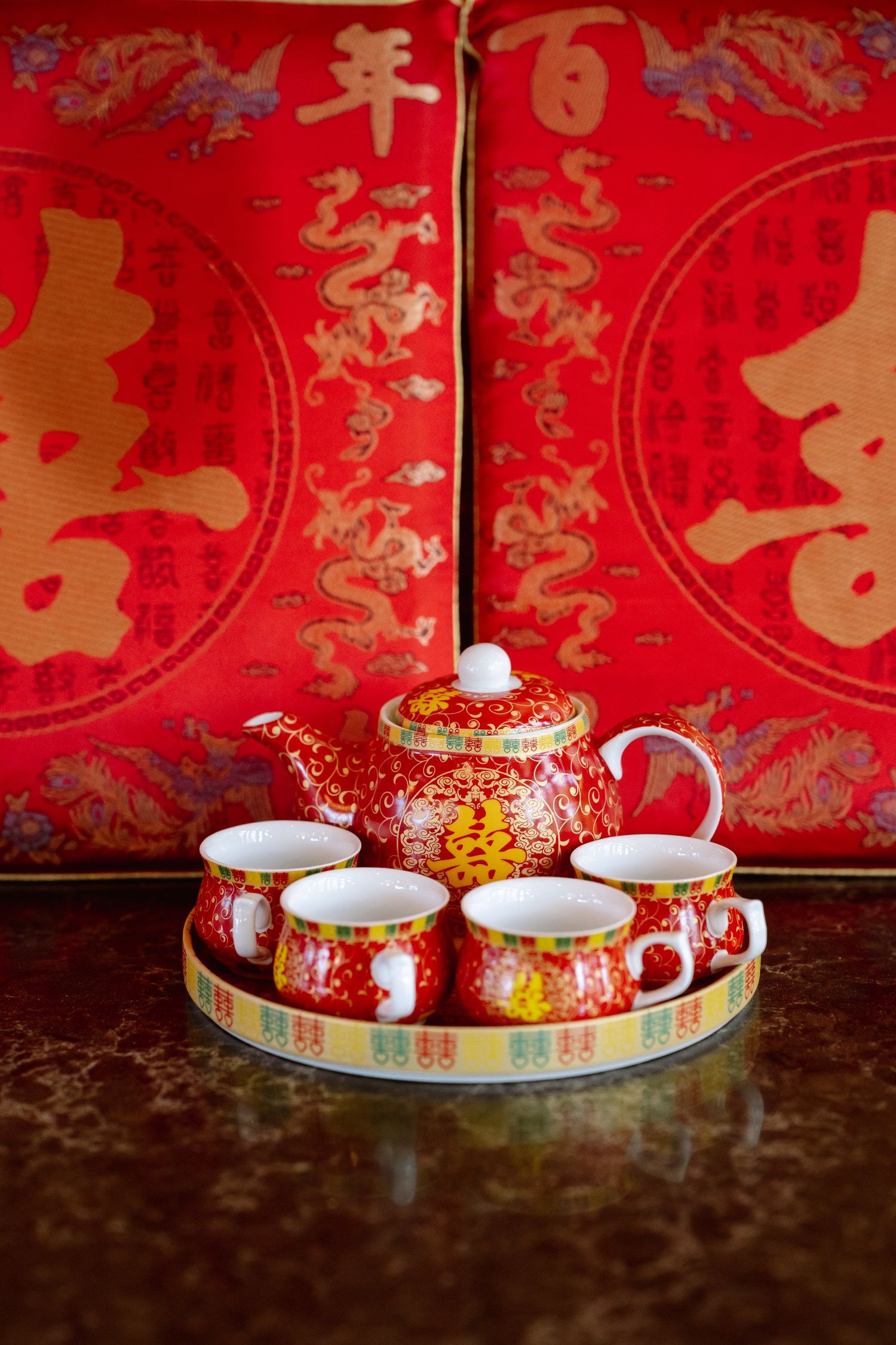 Red and gold Chinese wedding tea set with teapot and four cups on a tray, set before red cushions with dragon and double happiness designs.