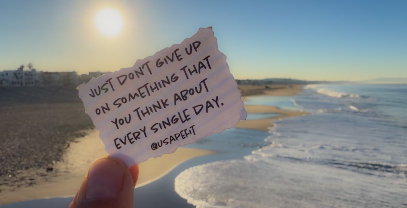 Hand holding a torn paper note with a motivational quote about not giving up, against a sunny beach, ocean waves and shoreline.