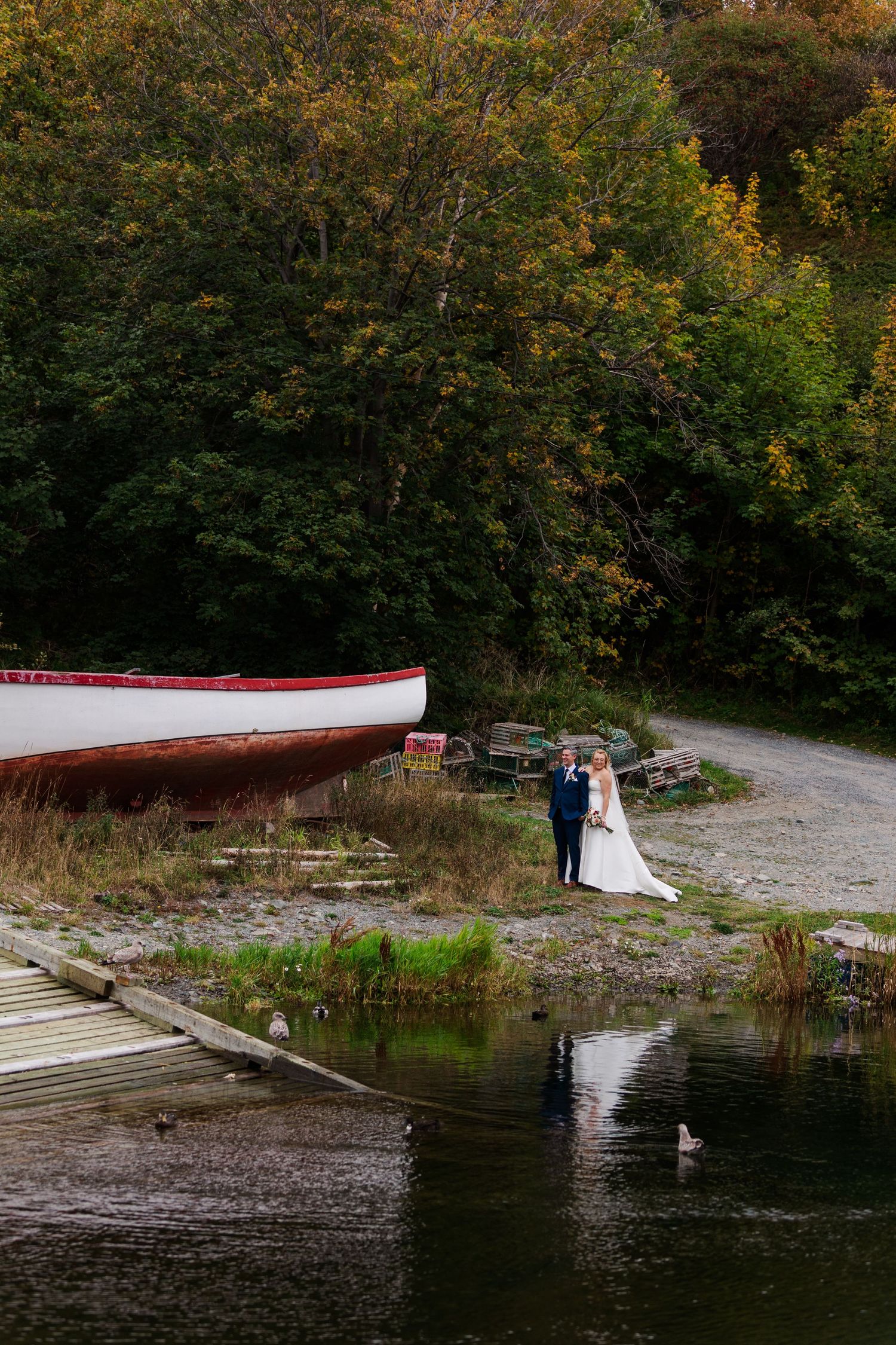 Bride and groom stand together on a rocky shore beside a red and white boat, with autumn trees and calm water in the background.