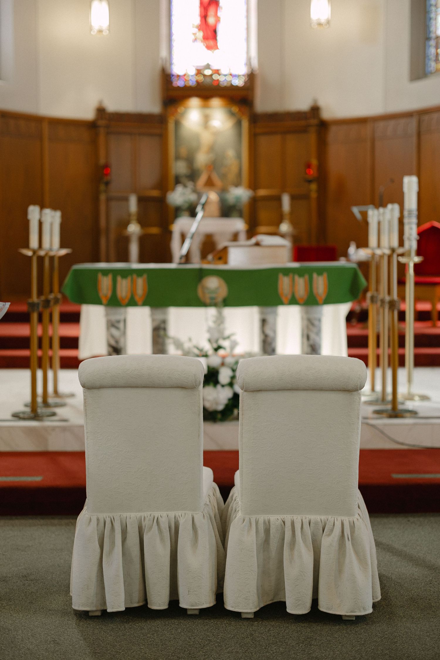 Two white slipcovered chairs face a green-draped church altar flanked by tall brass candlesticks and white floral arrangements.