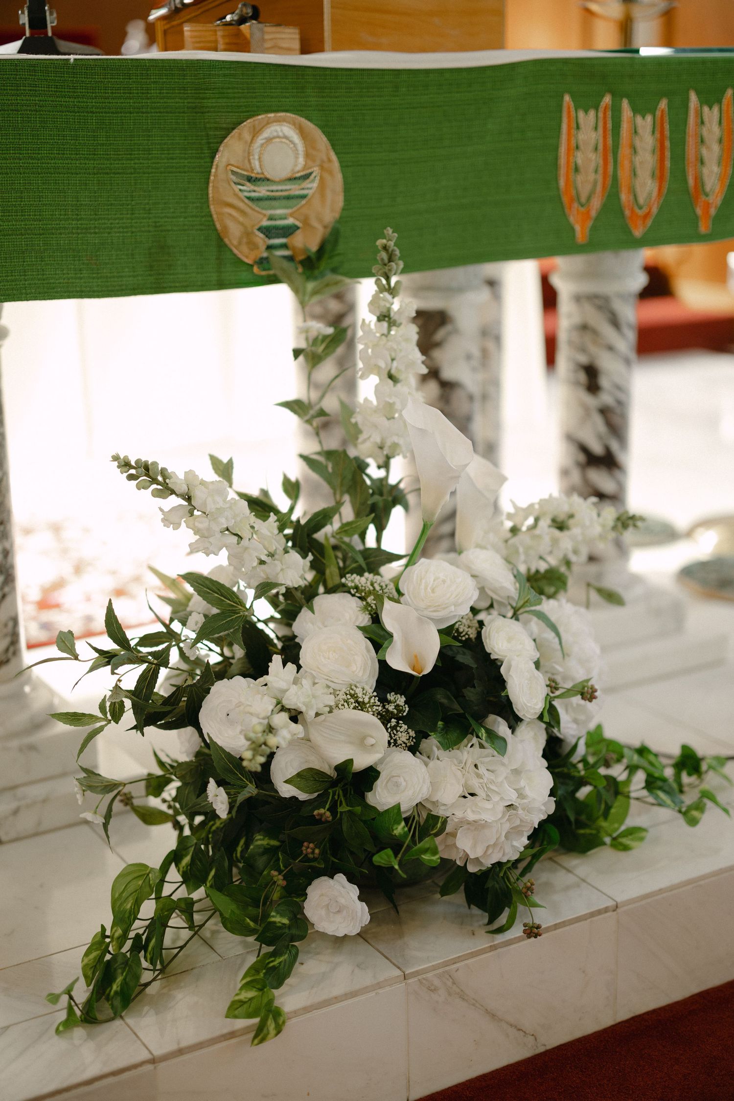 White roses, lilies, and trailing greenery rest on a marble altar step beneath a green cloth embroidered with a chalice emblem.