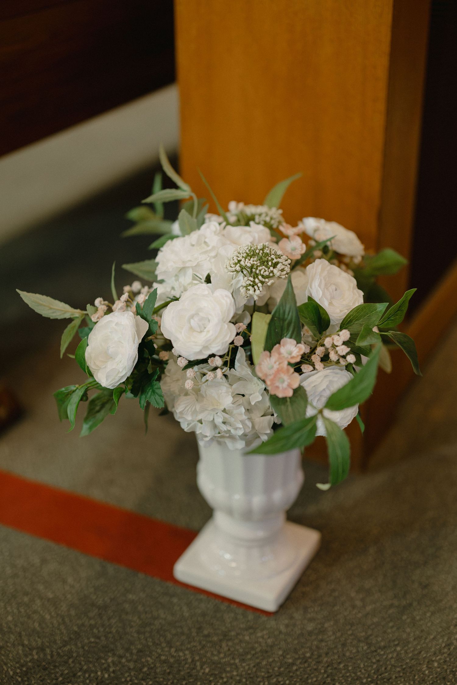 White roses, hydrangeas, and pink blooms with eucalyptus fill a white pedestal vase beside a wooden church pew.