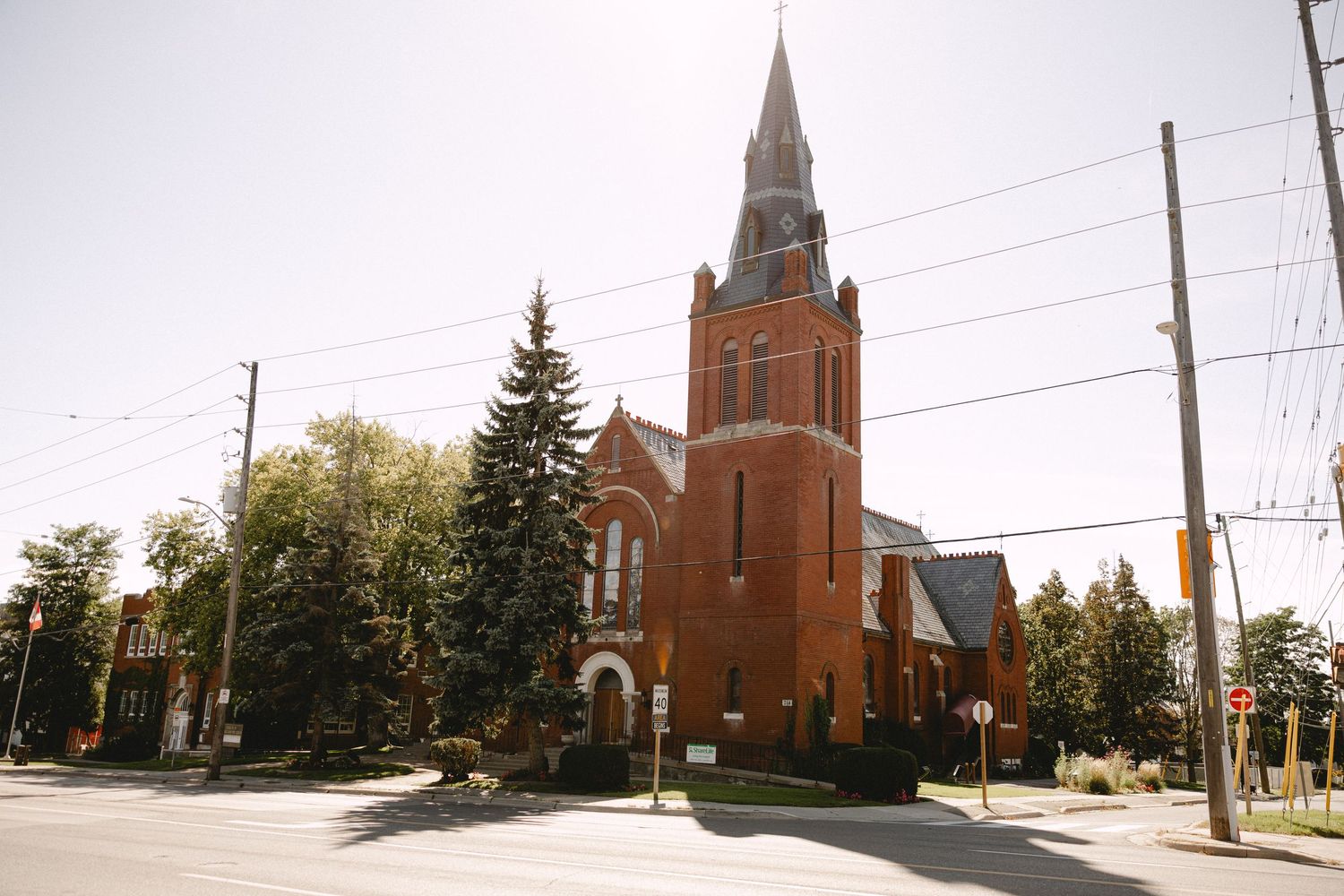Red brick church with a tall pointed steeple and evergreen trees on a sunny day along a quiet street.
