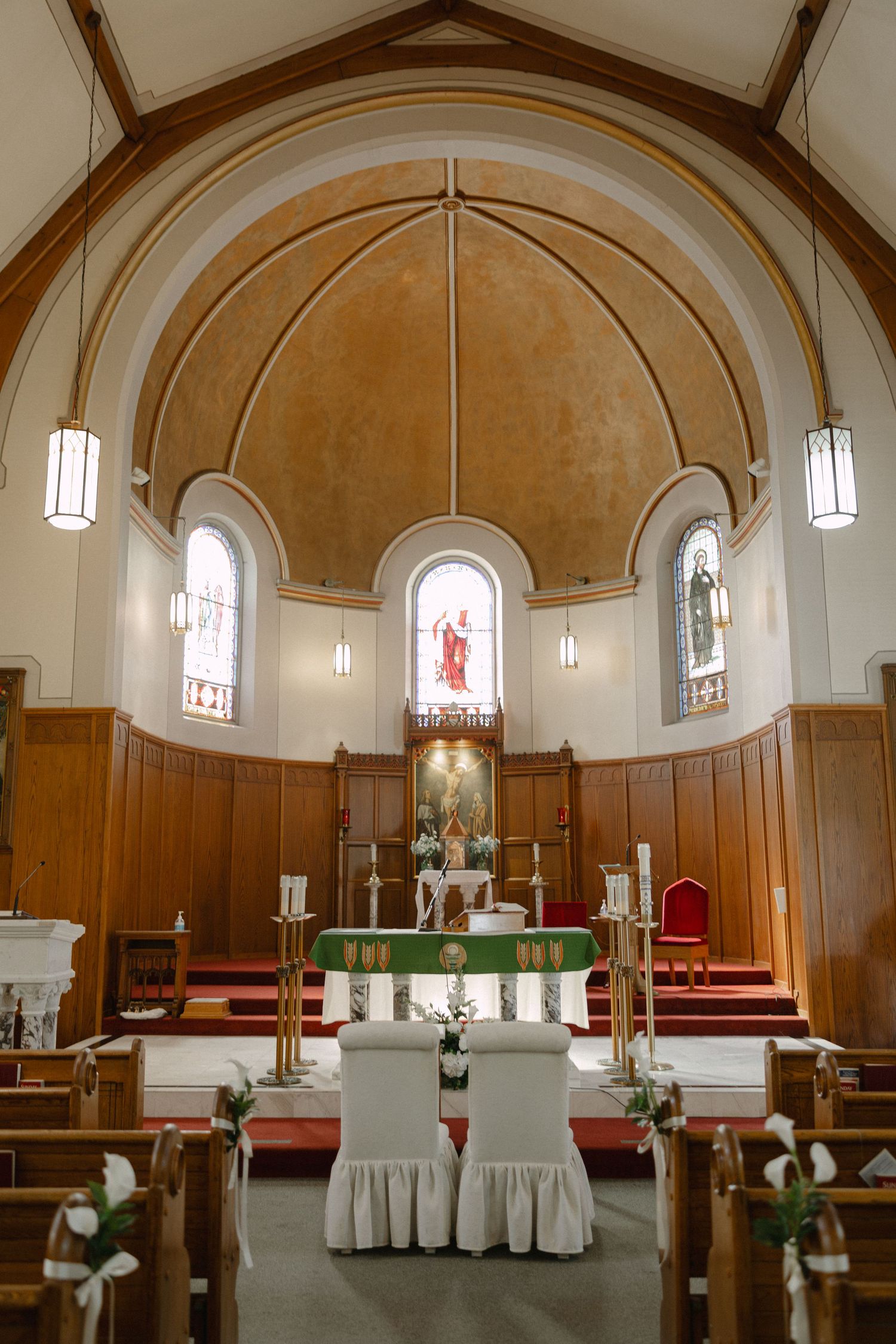 Church interior with wood paneling, arched ceiling, stained glass windows, and a green-draped altar with candles and white-covered chairs.