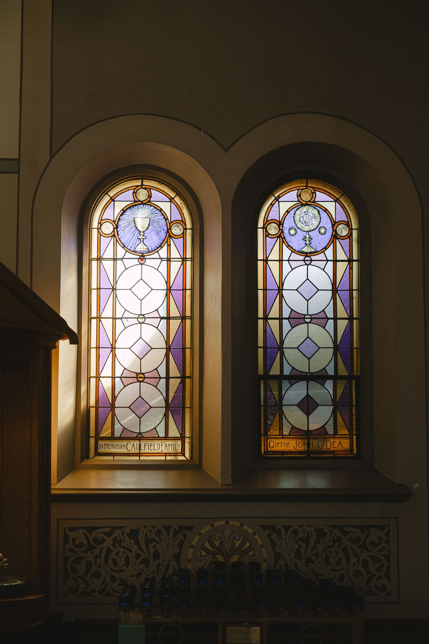 Two arched stained glass windows with purple and gold geometric patterns glow in a historic church interior.